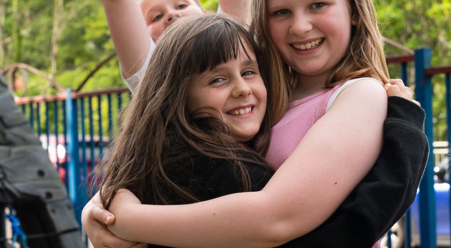 Two young girls hugging, with another child photo bombing in the background